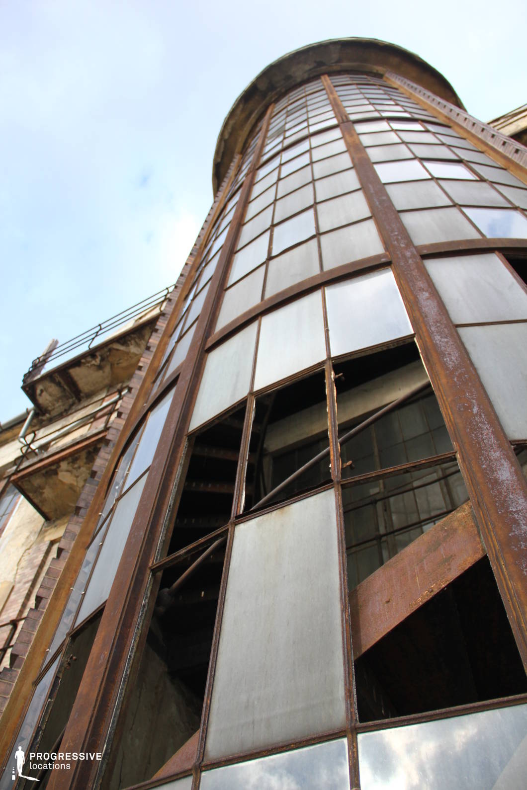 This image captures a towering, cylindrical glass stairwell structure with rusted metal framing, set against an aged brick facade. The perspective from below emphasizes the verticality and imposing nature of the architecture. Many glass panes are broken or missing, exposing the dark, skeletal remains of a spiral staircase inside, reinforcing a sense of abandonment and decay. The corroded surfaces and weathered materials create a gritty, industrial aesthetic reminiscent of old factories or institutional buildings. This location is well-suited for dystopian, post-apocalyptic, or urban exploration-themed films, offering a visually striking backdrop for tension-filled scenes, dramatic chases, or mystery-driven narratives.