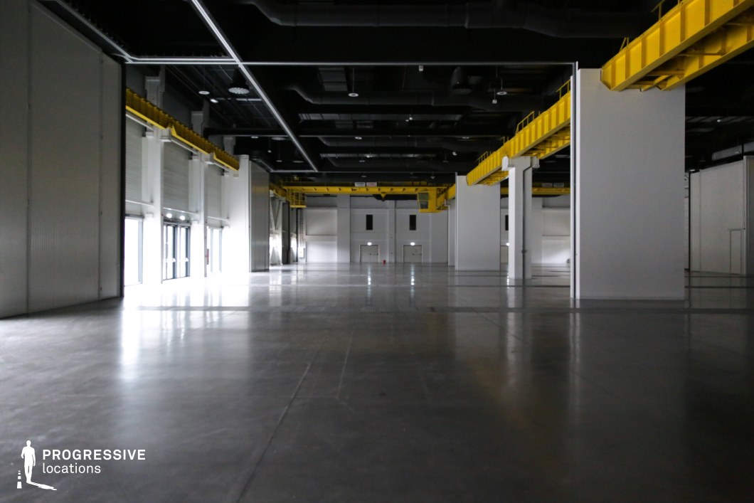 This vast industrial space features a polished concrete floor, high ceilings, and a structural grid of white columns supporting yellow overhead crane beams. The exposed black ceiling reveals ventilation ducts and industrial lighting, reinforcing the utilitarian aesthetic. Large roll-up doors allow natural light to enter, creating a dramatic contrast between illuminated and shadowed areas. The open floor plan and modular design make this location ideal for filming action sequences, chase scenes, or dystopian and sci-fi settings. The blend of industrial and futuristic elements provides flexibility for both high-tech and abandoned facility concepts.