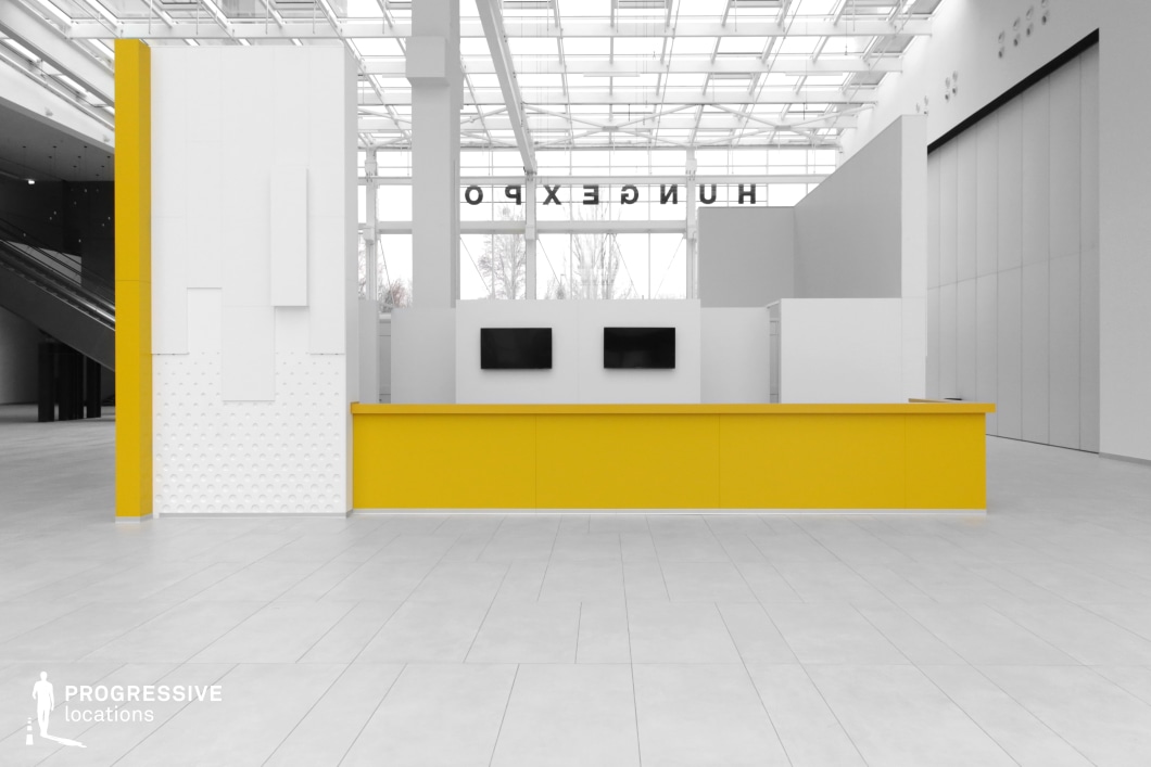 This spacious and contemporary exhibition hall features a sleek, minimalist design with a striking contrast between its neutral-toned environment and a bold yellow reception counter. The high ceiling, composed of a glass and metal framework, allows natural light to flood the area, enhancing its open and futuristic ambiance. Clean lines and geometric wall paneling contribute to a modern aesthetic, while the polished tile flooring reflects the structured, organized atmosphere of the space. The background features large partitions and escalators, suggesting a modular and multi-purpose layout. This location is ideal for filming corporate, futuristic, or high-tech scenes, as well as sequences set in airports, convention centers, or modern government facilities. Its neutral tones and open space make it adaptable for commercial shoots, product showcases, and cinematic productions requiring a sleek, professional setting.