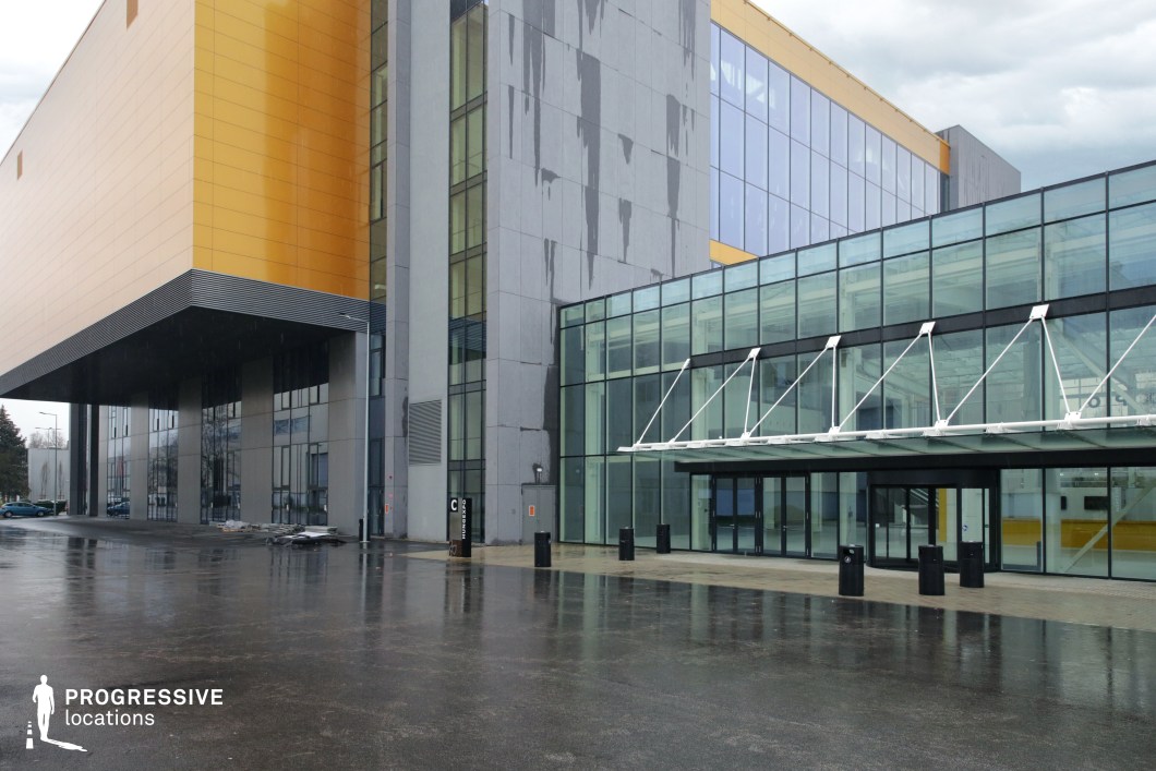 The image showcases a contemporary architectural structure with a striking combination of glass, metal, and concrete elements. The building features a bold, cantilevered golden panel that extends outward, creating a dynamic visual contrast against the sleek gray and glass facade. The entrance is marked by a transparent, steel-supported canopy, giving it an industrial yet sophisticated feel. The reflective surfaces amplify the modern aesthetic, while the large, open paved area in the foreground adds a sense of scale and accessibility. This location is ideal for filming corporate, futuristic, or thriller scenes, with its clean lines and industrial-modern design providing a visually compelling backdrop for high-tech environments, corporate power plays, or even dystopian urban settings.