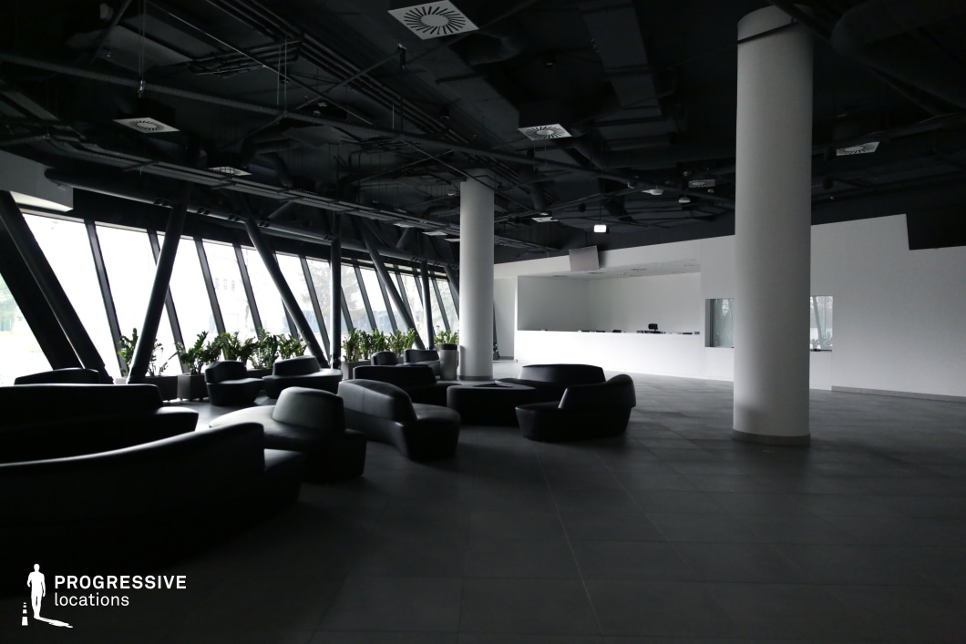 This modern lounge area features an open-plan design with sleek black furniture, floor-to-ceiling slanted glass windows, and exposed industrial-style ceiling elements. The contrast between the dark ceiling and light walls creates a sharp, contemporary aesthetic, while the white reception desk and cylindrical columns add a minimalist touch. The geometric window structures and subtle greenery contribute to a dynamic yet professional ambiance. This location is ideal for corporate, tech-thriller, or espionage scenes, serving as an airport lounge, futuristic office lobby, or high-end corporate waiting area.