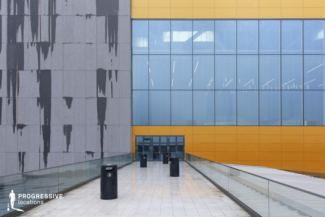 This striking urban setting features a juxtaposition of industrial and contemporary design elements. The walkway is flanked by a glass railing and leads to a set of glass doors framed by bold yellow paneling, creating a sharp contrast against the adjacent grey concrete wall with abstract black stains. The reflective glass windows mirror the surrounding structures, adding depth and an almost futuristic quality to the space. The structured pathway, lined with cylindrical black bollards, enhances the sense of direction and symmetry. This location is ideal for corporate thrillers, cyberpunk-style sci-fi, or high-stakes espionage scenes, offering a dynamic and visually engaging backdrop for characters entering or exiting a pivotal setting.