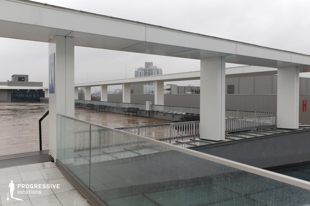 This expansive rooftop walkway features a sleek, modern design with clean white beams forming a structured, repetitive pattern. Glass railings and metal benches line the edges, reinforcing the contemporary aesthetic. The open space is framed by industrial-style buildings in the background, with a distant cityscape adding depth. The neutral tones and geometric lines create a minimalist, almost sterile atmosphere. This location is ideal for filming corporate, espionage, or futuristic scenes, serving as a rooftop meeting point, chase sequence setting, or high-tech surveillance post. Its structured layout provides opportunities for dynamic camera movements and dramatic lighting contrasts.