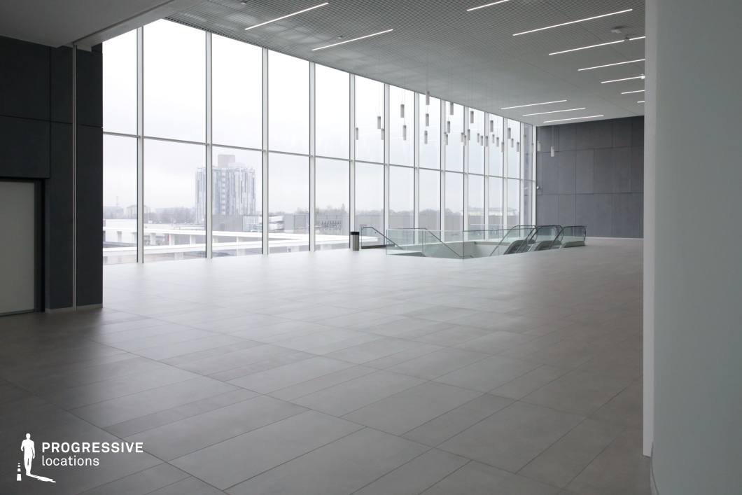 This image presents a vast, open interior space with a sleek, minimalist design. Dominated by expansive floor-to-ceiling windows, the area is bathed in natural light, offering a panoramic view of the surrounding cityscape. The high ceiling is adorned with linear recessed lighting and suspended cylindrical fixtures, adding a refined, contemporary touch. The polished tiled floor and neutral color palette enhance the clean, corporate aesthetic. The open floor plan, along with a centrally positioned escalator, suggests a transportation hub or high-end commercial setting. This location is ideal for filming corporate dramas, sci-fi sequences, high-stakes negotiations, or futuristic settings, providing an elegant yet neutral backdrop that allows for versatile storytelling.