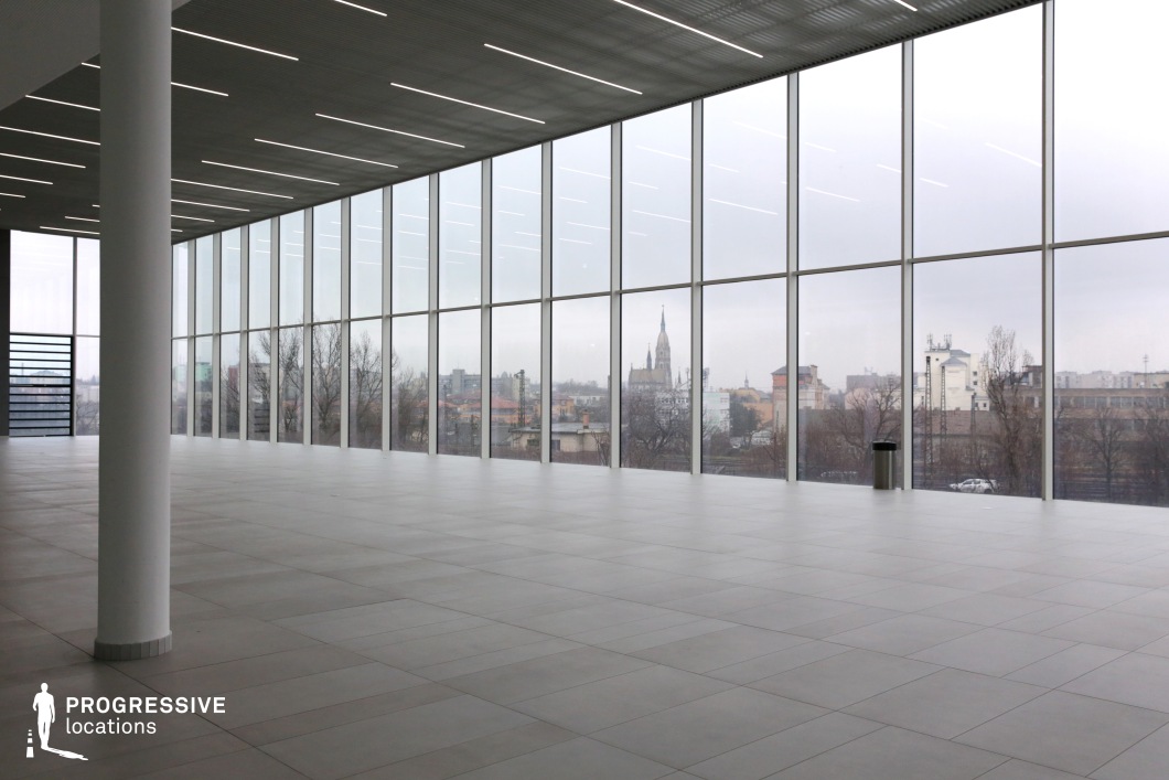 This expansive, modern interior features floor-to-ceiling glass windows that provide an unobstructed panoramic view of the surrounding cityscape, including historic architecture and contemporary structures. The minimalistic design, with its large, light-colored tiled flooring, sleek ceiling lighting, and structural white columns, creates a clean and spacious environment with a professional and futuristic aesthetic. The location is ideal for filming corporate scenes, high-end fashion shoots, or futuristic narratives, offering a versatile backdrop for business meetings, technology-focused storytelling, or moments of introspection set against an urban skyline.