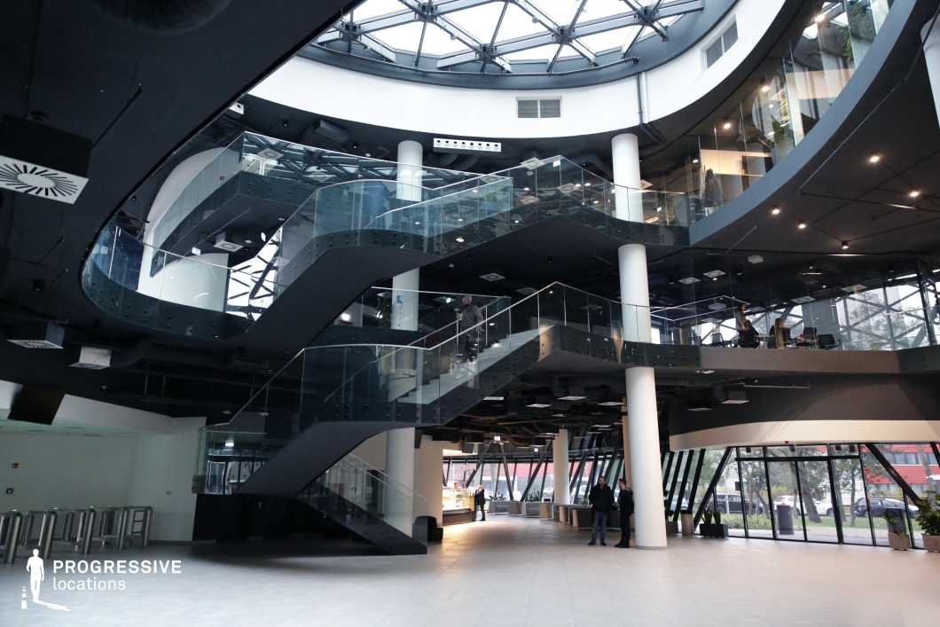 This striking multi-level interior features a spacious atrium with a futuristic aesthetic, dominated by sleek black staircases with transparent glass railings that zigzag between floors. The space is framed by curved balconies with glass partitions, creating a dynamic interplay of light and structure. The ceiling is composed of a geometric glass dome, allowing natural light to enhance the high-contrast monochromatic palette. White cylindrical columns support the open-plan structure, adding to the modern and sophisticated design. This location is ideal for corporate, cyber-thriller, or high-tech espionage films, serving as a contemporary office, high-security facility, or futuristic research hub. Its clean architectural lines and reflective surfaces offer excellent opportunities for dramatic cinematography, emphasizing movement and verticality.
