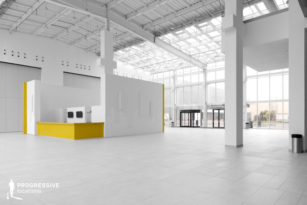 This expansive, modern atrium features a sleek, open-concept design with high ceilings and large glass walls, allowing for ample natural light. The dominant white color scheme, combined with clean lines and geometric structures, enhances the space's minimalist and industrial aesthetic. A standout element is the centrally placed white structure with a bold yellow counter, adding a subtle contrast to the otherwise neutral environment. The polished tile flooring and exposed ceiling framework contribute to a contemporary and slightly futuristic atmosphere. This location is ideal for filming corporate settings, futuristic or sci-fi environments, or high-end commercial spaces. Its open layout makes it suitable for large-scale cinematic shots, establishing scenes, or dynamic tracking sequences.