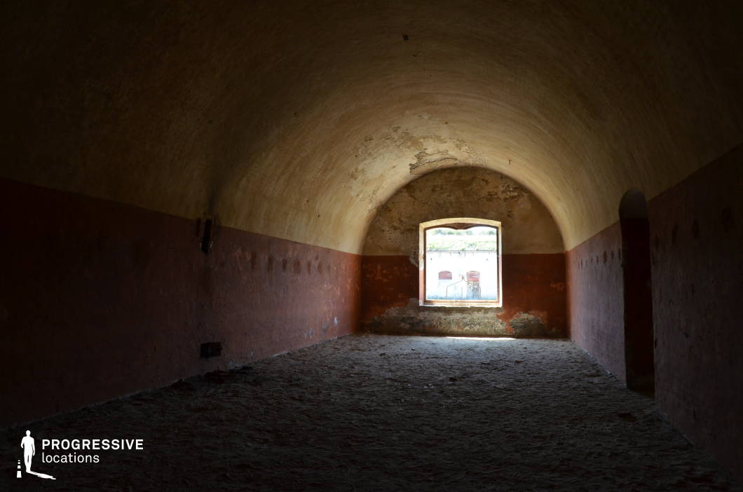 The image features a dimly lit, arched chamber with a worn, textured ceiling and faded red lower walls, evoking a sense of history and abandonment. The uneven floor and peeling paint enhance the aged and rugged atmosphere. A single, small window serves as the only light source, casting a soft glow that highlights the rough, decayed surfaces and provides a glimpse of the outside world. The space has a confined yet imposing presence, making it ideal for filming historical, war, or mystery films. This location could serve as an old prison cell, secret meeting room, or wartime hideout, adding depth to tense and dramatic storytelling.