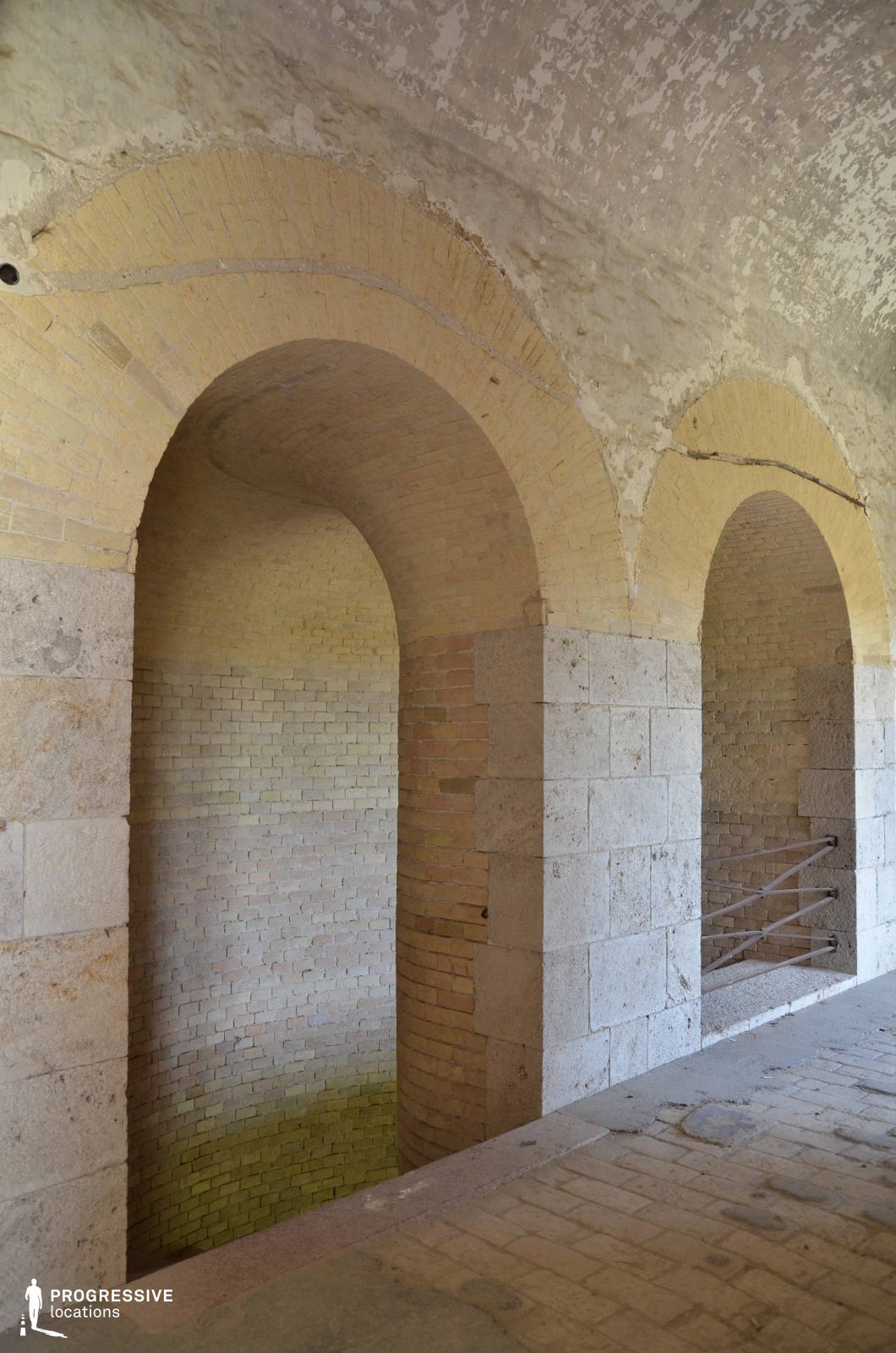 This aged architectural space features a series of large, rounded brick archways leading into shadowy alcoves, framed by robust stone columns. The weathered surfaces, with peeling plaster and exposed masonry, add a sense of historical depth, while the muted earth tones of the brickwork enhance the structure’s timeless appeal. The interplay of light and shadow emphasizes the depth of the passage, creating a strong visual rhythm. This location is ideal for historical dramas, period films, or suspenseful scenes requiring an old-world, fortress-like atmosphere, making it a perfect setting for moments of intrigue, secret meetings, or contemplative character-driven storytelling.