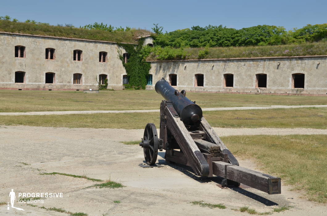 This well-preserved military fortress features a long, stone-built structure with arched window openings and vegetation creeping over parts of the walls. The foreground is dominated by an aged black cannon mounted on a weathered wooden carriage, reinforcing the historical and defensive character of the site. The open courtyard, with grass and dirt pathways, enhances the sense of a functional military installation from a bygone era. This location is ideal for historical war films, period dramas, or adventure stories, providing an authentic backdrop for battle reenactments, military strategies, or exploration scenes set in the 18th or 19th century.
