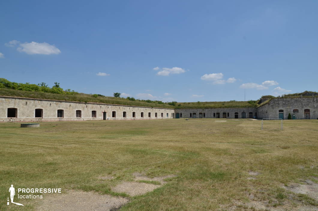 This open, fortress-like structure features a long, low stone building with multiple evenly spaced doorways, surrounding a vast grassy courtyard. The weathered stone walls and partially overgrown rooftop blend with the natural environment, suggesting an old military installation or historical barracks. The absence of modern elements enhances the location’s timeless and slightly desolate character. The wide, open space provides an excellent backdrop for historical battle reenactments, war dramas, or dystopian and post-apocalyptic settings. The fort’s architecture also lends itself to strategic and suspenseful sequences, such as military planning scenes, last-stand confrontations, or even secretive underground hideout narratives.