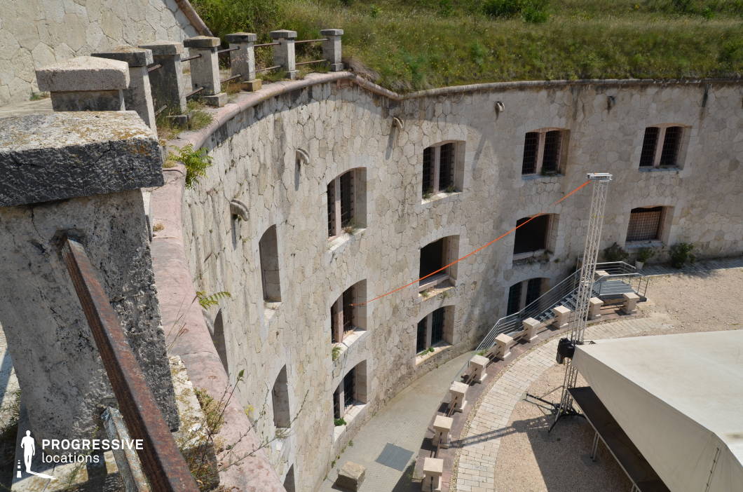 The image showcases a circular stone fortress with a multi-level courtyard, constructed from weathered limestone blocks. The exterior walls are lined with evenly spaced, arched windows fitted with iron bars, reinforcing a historic or defensive architectural style. A curved stone railing with aged metal reinforcements surrounds the upper perimeter, overlooking the central open space below. The courtyard features a semi-circular arrangement of stone benches, a modern staircase, and a temporary canopy structure, blending historical and contemporary elements. The partially overgrown embankment above adds to the aged atmosphere. This location is well-suited for filming historical dramas, medieval or military period pieces, as well as dystopian or prison-themed narratives, offering a dramatic backdrop for tense confrontations, escape sequences, or strategic discussions.