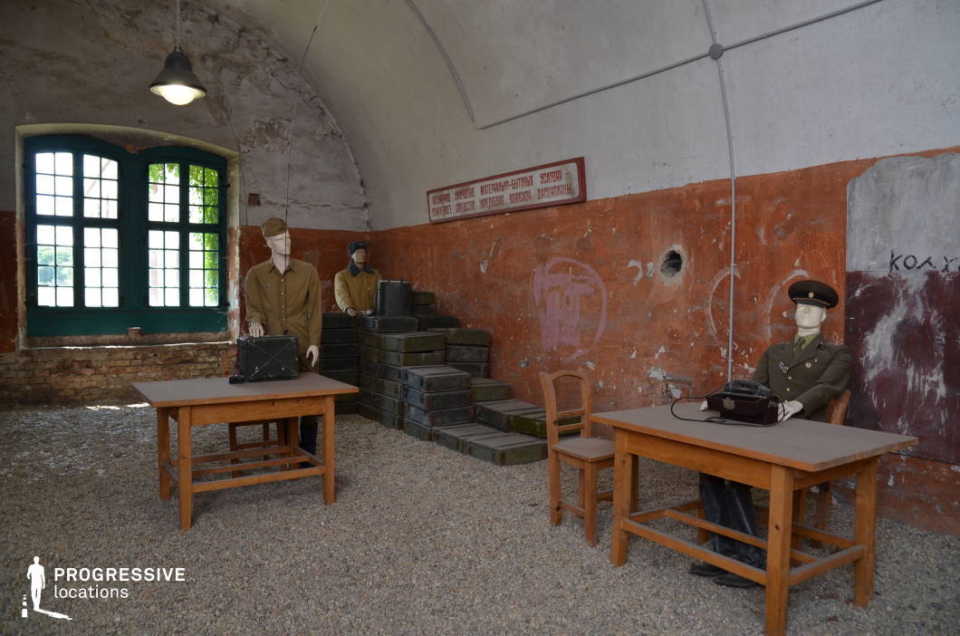 This rustic military-themed room is housed within an arched, aged structure with exposed brickwork and peeling paint on the walls, evoking a historical or Cold War-era setting. The distressed orange and white walls, combined with the rough gravel floor, enhance the atmosphere of a repurposed bunker or wartime operations center. Simple wooden tables and chairs, along with stacked military storage boxes, reinforce the utilitarian nature of the space. The large, multi-paned window with a deep green frame allows natural light to filter in, contrasting the otherwise dim and enclosed atmosphere. This location is ideal for filming historical, war-themed, or espionage scenes, offering a visually compelling backdrop for military strategy discussions, underground resistance operations, or abandoned outpost settings.