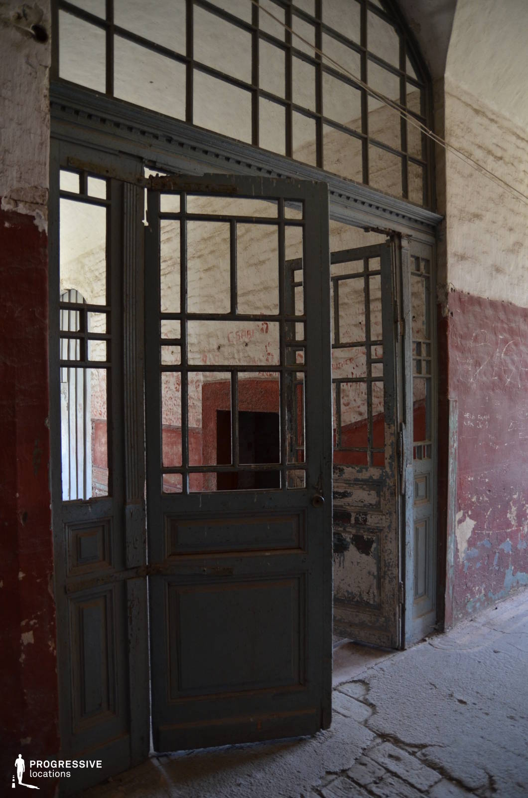 This image presents a set of tall, metal-framed doors with intricate glass paneling, partially ajar, leading into a worn, abandoned space. The faded blue paint is chipped, revealing layers of history, while the surrounding walls display exposed brick, peeling plaster, and remnants of old markings. Above, a large arched transom window allows light to filter through, enhancing the atmosphere of neglect and faded grandeur. The cracked and uneven floor further adds to the sense of decay. This location is ideal for filming historical dramas, psychological thrillers, or mystery narratives, offering a visually rich setting for themes of abandonment, secrecy, or the passage of time.