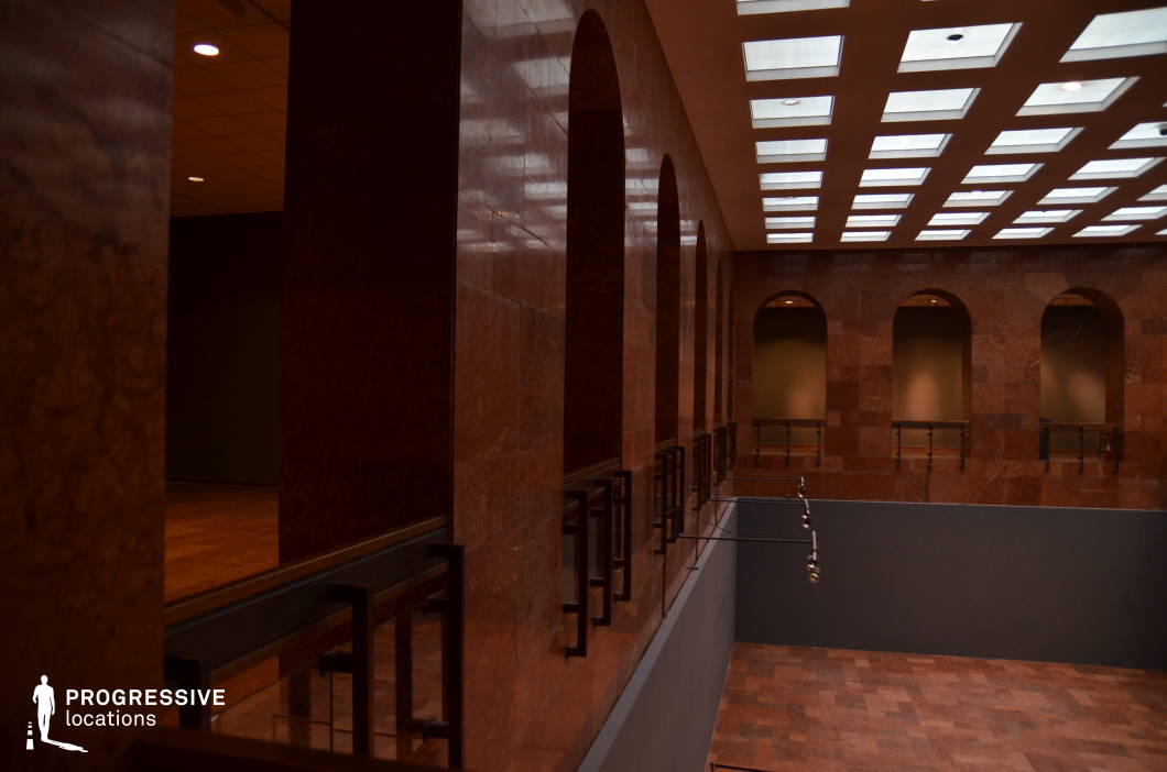 This sophisticated interior space is defined by its rich, warm-toned stone walls and flooring, evoking a classical yet modern ambiance. The symmetrical series of large arched openings on both levels, framed by sleek metal railings, adds a sense of grandeur and depth. The ceiling is adorned with a geometric grid of recessed lighting panels, casting a soft, diffused illumination that enhances the structured aesthetic. The interplay of shadow and light across the polished stone surfaces adds texture and atmosphere. This location is ideal for historical dramas, corporate thrillers, or mystery films, offering a refined, imposing backdrop suitable for intense dialogues, confrontational meetings, or high-stakes corporate intrigue.