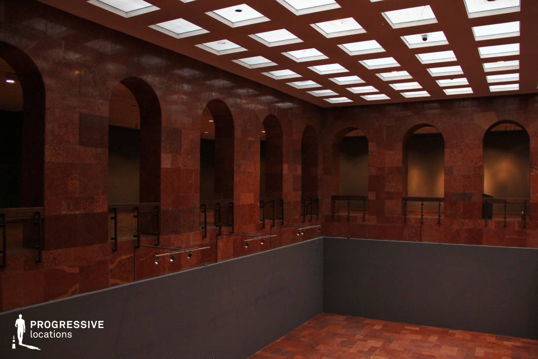 This richly textured interior features warm-toned stone walls adorned with a series of evenly spaced, grand arches, creating a classical yet contemporary atmosphere. The ceiling incorporates a geometric grid of recessed lighting, casting an ambient glow over the space. A mezzanine level with glass and metal railings overlooks a sunken central area, adding depth and perspective. The architectural balance of symmetry and earthy materials makes this an ideal filming location for historical dramas, high-end corporate settings, or sophisticated thriller scenes. The play of light and shadow within the arches enhances its cinematic potential, offering dramatic framing opportunities.