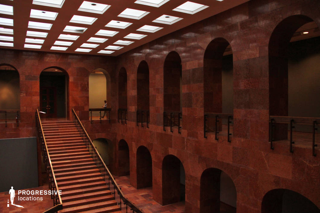 This imposing interior features a grand staircase leading to an upper-level gallery framed by a series of evenly spaced, rounded arches. The entire space is clad in rich red marble, giving it a stately, institutional feel. The ceiling consists of a grid of recessed rectangular lighting panels, casting a diffused glow that enhances the hall’s dramatic ambiance. Dark metal railings contrast with the warm-toned stone, adding a contemporary touch to the classical architectural elements. This location is well-suited for historical, political, or mystery films, serving as a courthouse, museum, or government building. The symmetrical design and strong vertical lines provide a powerful visual composition for cinematic storytelling.