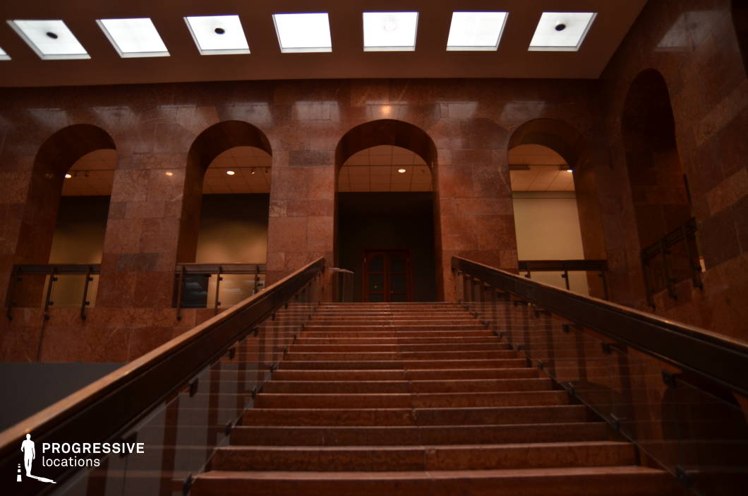 This imposing staircase, clad in polished brown marble, leads to a series of arched openings, exuding a classical yet institutional grandeur. The warm, earthy tones of the stone contrast with the sleek glass railings, blending tradition with modernity. Overhead, rectangular skylights allow diffused lighting to emphasize the depth and symmetry of the space. The repetition of arches enhances the location’s formal and somewhat enigmatic atmosphere. This setting is well-suited for filming historical dramas, political thrillers, or mystery narratives, functioning as a courthouse, museum, or secretive government facility. The stairway’s strong perspective also makes it ideal for dynamic tracking shots and dramatic character entrances.