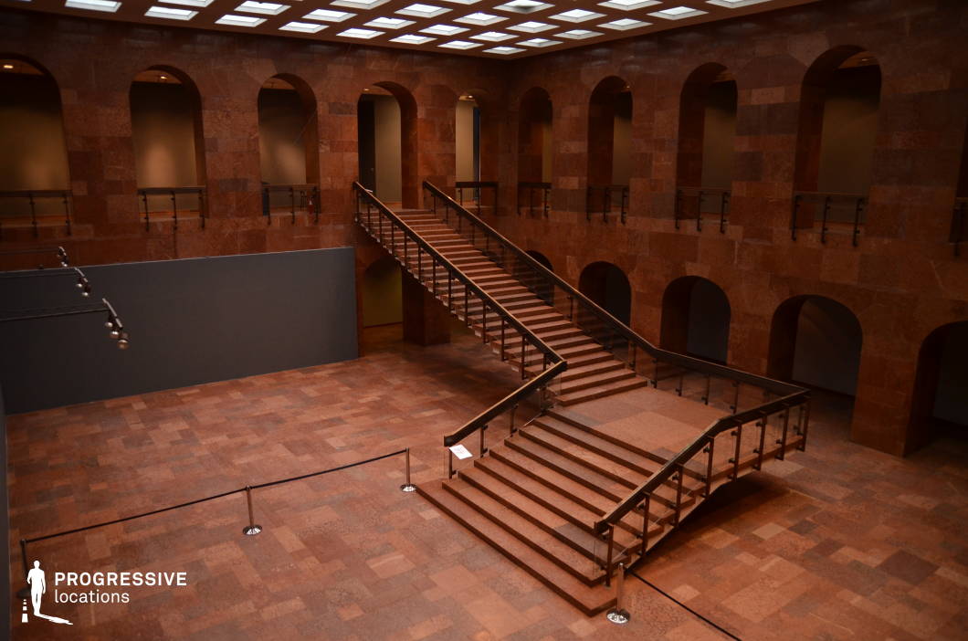 This imposing interior space is defined by its deep red marble surfaces, creating a rich and dramatic atmosphere. A grand central staircase with symmetrical flights dominates the hall, leading to an upper-level gallery lined with evenly spaced arched openings. The ceiling features a grid of recessed lighting, adding a modern contrast to the historic architectural feel. The vast open floor space and enclosed upper level provide a sense of depth and scale, making this an ideal setting for filming period dramas, mystery thrillers, or high-society power struggles. The strong geometric design, luxurious materials, and monumental presence make it suitable for scenes requiring an opulent, formal, or institutionally significant backdrop.