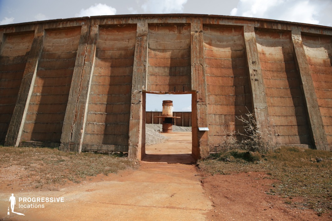 This massive circular concrete structure, marked by weathered surfaces and rust-colored staining, evokes a sense of decay and forgotten industry. The thick, reinforced walls are divided into vertical sections with deep-set supports, creating a rhythm of shadow and texture. A small, rectangular doorway punctuates the structure, framing a distant rusted cylindrical chimney and additional industrial elements in the background. The cracked surfaces and overgrown edges contribute to a dystopian, post-apocalyptic aesthetic. This location is ideal for filming scenes set in abandoned industrial zones, science-fiction wastelands, or historical narratives centered around defunct infrastructure. The combination of heavy brutalist architecture and desolate surroundings makes it particularly suited for thriller, war, or survival-themed productions.