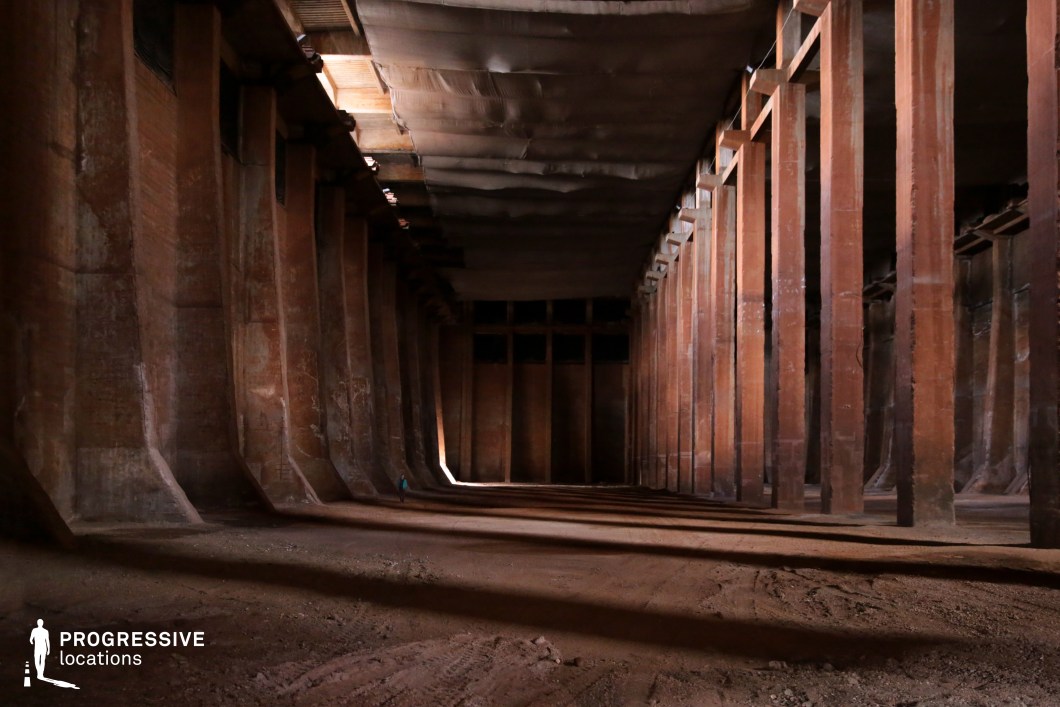 This cavernous space features towering, evenly spaced concrete pillars supporting a heavy industrial ceiling, draped with aged fabric that diffuses the dim ambient light. The reddish-brown surfaces of the walls and columns create a raw, monolithic atmosphere, while the dirt-covered ground adds to the abandoned, subterranean feel. The linear perspective of the columns and deep shadows enhance the space’s sense of scale and mystery. This location is perfect for filming underground hideouts, secret industrial facilities, or dystopian survival scenes, offering a dramatic and immersive setting for high-stakes cinematic moments.