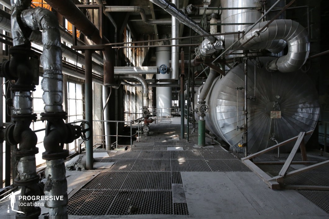 This expansive industrial space features an intricate network of pipes, valves, and large cylindrical tanks, creating a raw and mechanical atmosphere. The aging metal surfaces, covered in dust and rust, contribute to a worn, abandoned feel, while the grated metal flooring and overhead piping add layers of depth and texture. The large, multi-paned windows allow streaks of natural light to filter in, casting dramatic shadows across the industrial elements. The combination of metallic tones, faded paint, and exposed framework makes this location ideal for filming dystopian, cyberpunk, or thriller scenes. Its maze-like structure and mechanical complexity offer a compelling setting for factory-based action sequences, sci-fi laboratory interiors, or espionage thrillers requiring an industrial hideout or high-tension confrontation space.