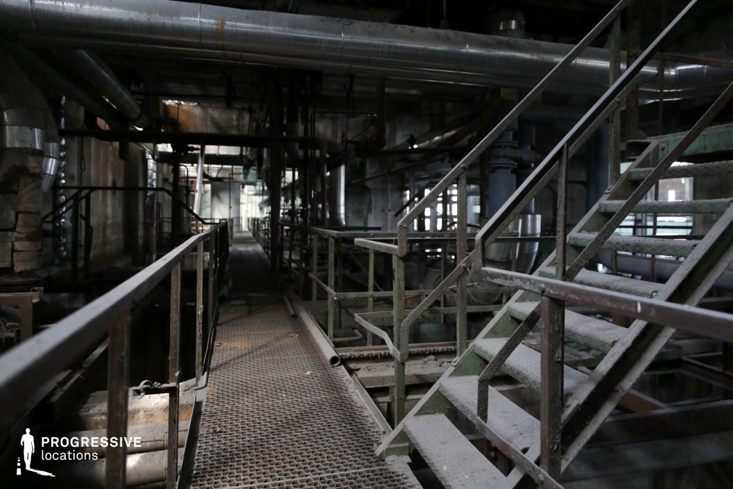 This decayed industrial setting features a network of rusted metal staircases, grated walkways, and an overhead tangle of pipes, creating a dense, mechanical environment. The darkened interior, layered with dust and corrosion, enhances the abandoned and hazardous aesthetic. Exposed beams and industrial tubing dominate the ceiling, while the long, narrow pathways add a sense of depth and unease. This location is highly suited for filming dystopian, action, or thriller sequences, particularly chase scenes, secretive meetings, or high-stakes confrontations in a post-apocalyptic or cyberpunk setting.