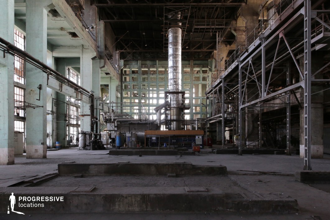 This vast, abandoned industrial hall features towering metal frameworks, peeling pastel-green concrete walls, and a massive central machine with an imposing metallic chimney structure. The high ceilings and large, gridded windows allow diffused light to cast long shadows, enhancing the decayed, utilitarian atmosphere. Rusted pipes, exposed steel beams, and scattered remnants of machinery contribute to the sense of a once-active space now left to time. The setting is ideal for dystopian, cyberpunk, or post-apocalyptic film scenes, as well as action sequences, clandestine meetings, or factory-set thrillers where a gritty, industrial aesthetic is essential.