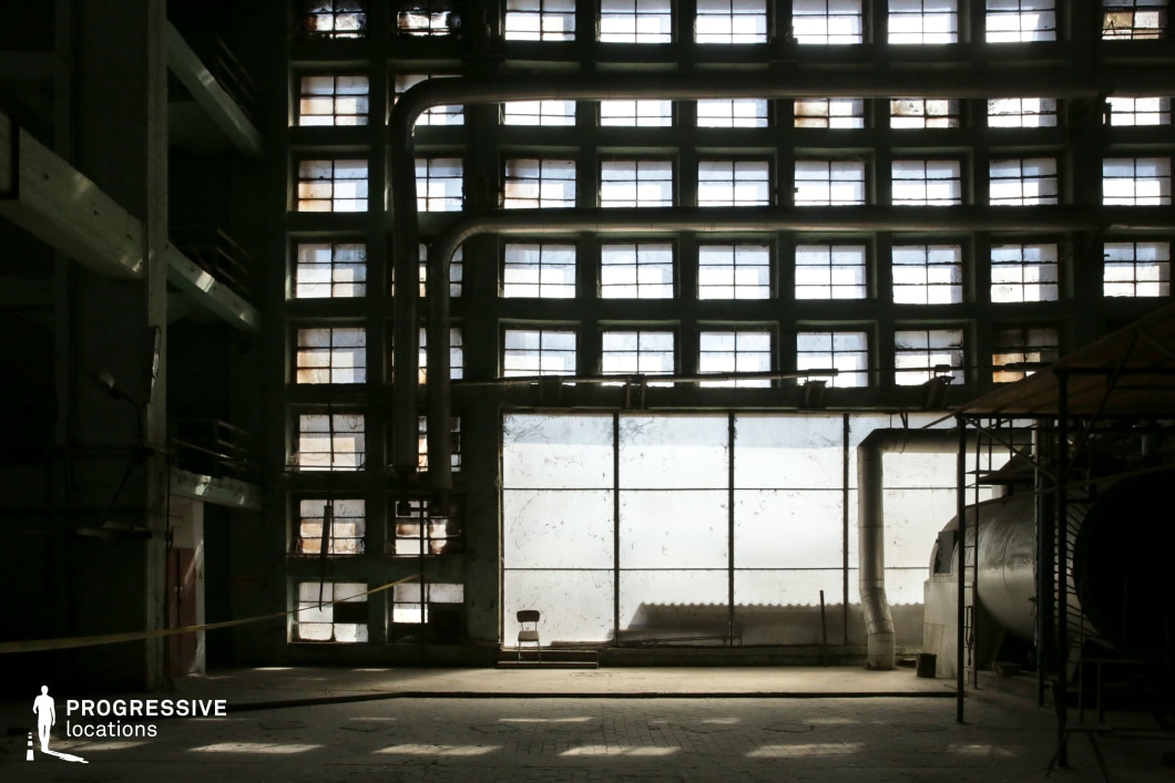 This vast, decaying industrial hall is dominated by a grid of large, dirty windows that filter in dim, diffused light, casting dramatic geometric shadows on the worn concrete floor. The structure features towering metal pipes running along the walls, a rusted cylindrical tank, and remnants of old machinery, emphasizing its industrial past. The imposing height of the space and the repetitive window pattern create a sense of isolation and emptiness. The mix of grime, dust, and structural decay gives the location a haunting, post-apocalyptic aesthetic. Ideal for thriller, dystopian, or crime films, this setting works well as an abandoned factory, secret research facility, or a place for clandestine meetings. The interplay of light and shadow enhances the visual drama, making it perfect for atmospheric cinematography.