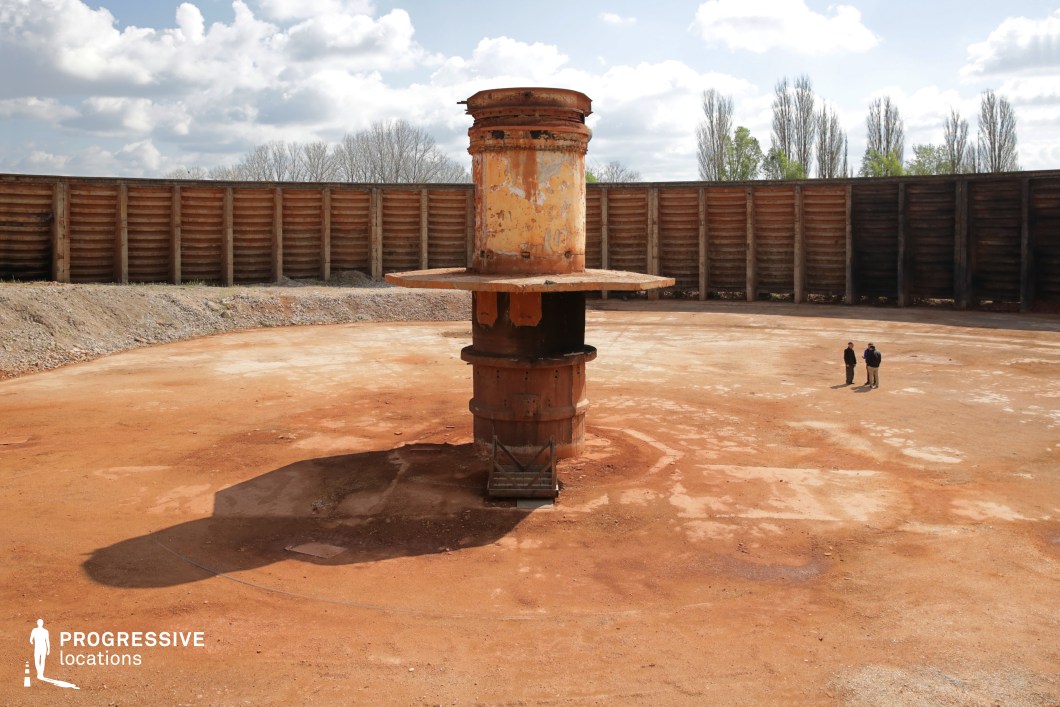 This vast circular enclosure, surrounded by high wooden retaining walls, creates a stark and isolated atmosphere reminiscent of a dystopian or post-apocalyptic setting. The rusted central metal structure, weathered and corroded, stands as the focal point in the middle of the reddish, compacted earth, evoking a sense of abandonment and decay. The open expanse and lack of modern infrastructure enhance the desolate and industrial aesthetic. The contrast between the rigid wooden walls and the worn metal component adds to the location’s raw, utilitarian feel. This setting is ideal for sci-fi, dystopian, or action film productions, offering a dramatic and enclosed space suitable for intense confrontations, experimental tests, or clandestine meetings in a world that feels disconnected from reality.