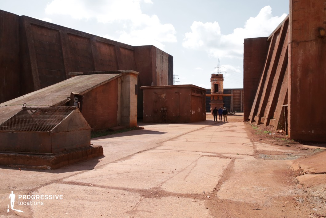 This rugged industrial location is dominated by massive, weathered concrete walls with a reddish-brown patina, giving the space a raw, aged appearance. The geometric structures, reinforced buttresses, and angular pathways create a fortress-like atmosphere. The cracked pavement and exposed metal elements contribute to the feeling of abandonment and decay. A lone tower-like structure in the background adds vertical contrast to the otherwise enclosed space. This location is ideal for filming dystopian, post-apocalyptic, or action-oriented scenes, serving as a military compound, prison yard, or secret industrial facility. The vast open areas and strong architectural lines provide dynamic framing opportunities for dramatic sequences.