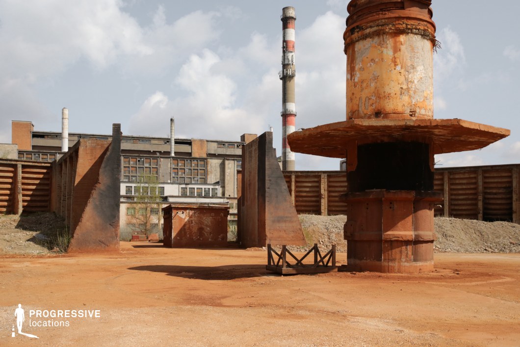 This vast industrial site features rusted metal structures, decayed machinery, and weathered concrete walls, creating a raw, post-apocalyptic atmosphere. The towering smokestacks and broken-down architectural elements evoke a sense of abandonment and decay, while the reddish-brown dirt ground enhances the rugged, dystopian aesthetic. The layered composition, with the factory complex in the background and remnants of old industrial equipment in the foreground, provides a dramatic visual depth. This location is ideal for dystopian, sci-fi, or action films, serving as a backdrop for chase sequences, secret experiments, or post-collapse urban survival settings.