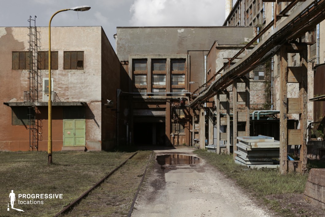 The image depicts a decayed industrial complex with rusted metal structures, cracked concrete walls, and weathered brick facades. A set of narrow railway tracks leads toward a shadowy entrance, adding depth and perspective to the scene. Overhead pipelines and corroded support beams create a layered, mechanical aesthetic, reinforcing the location's industrial past. The muted color palette of faded browns, grays, and rusted oranges contributes to a gritty, post-apocalyptic atmosphere. This location is ideal for filming dystopian, thriller, or crime scenes, offering a visually rich backdrop for abandoned facility sequences, industrial espionage, or chase scenes through derelict infrastructure.