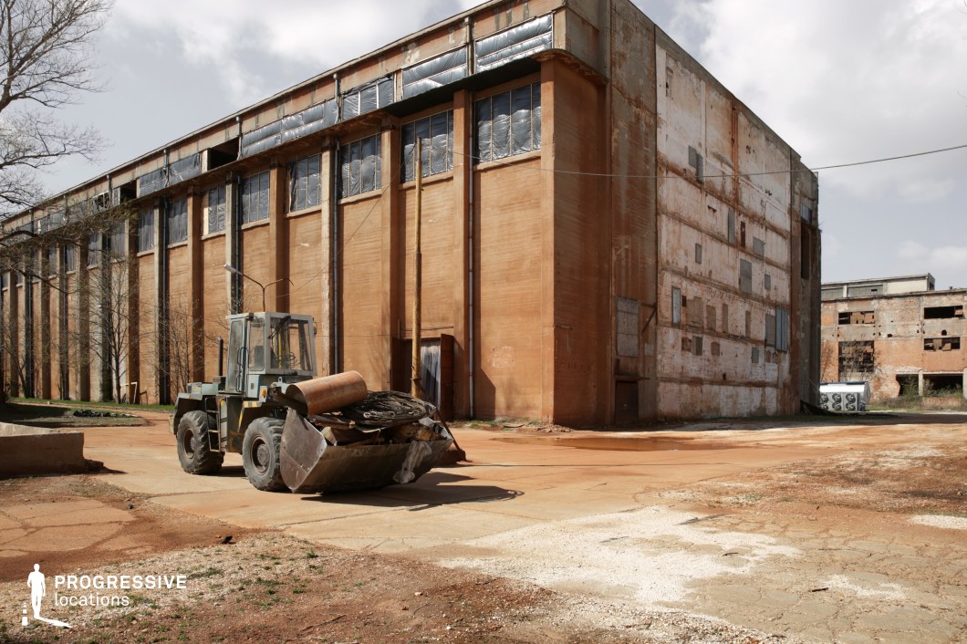 This decayed industrial complex features towering, rust-colored concrete walls with peeling paint and broken windows, creating a sense of neglect and abandonment. The structure's vertical columns and repetitive window patterns emphasize its massive scale, while the boarded-up sections suggest a long-forgotten past. The cracked ground and scattered debris enhance the raw, gritty atmosphere. A large, rusted machine sits in the foreground, reinforcing the industrial decay. This location is ideal for filming dystopian, post-apocalyptic, or crime-oriented scenes, serving as an abandoned factory, secret hideout, or derelict urban setting. The interplay of light and shadow through the broken windows adds visual depth, perfect for high-contrast cinematography.