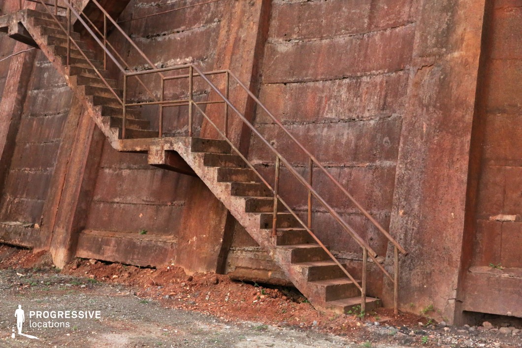 This weathered industrial staircase, constructed of rusted metal and concrete steps, is attached to a massive red-toned concrete wall that exhibits visible wear, cracks, and layers of corrosion. The rough, aged textures of the structure contrast with the stark geometric form of the staircase, creating a gritty and raw visual aesthetic. The surrounding ground, composed of loose dirt and debris, adds to the sense of abandonment and decay. This location is ideal for filming scenes in the post-apocalyptic, crime, or thriller genres, offering a striking backdrop for chase sequences, clandestine meetings, or tense confrontations in an industrial setting.