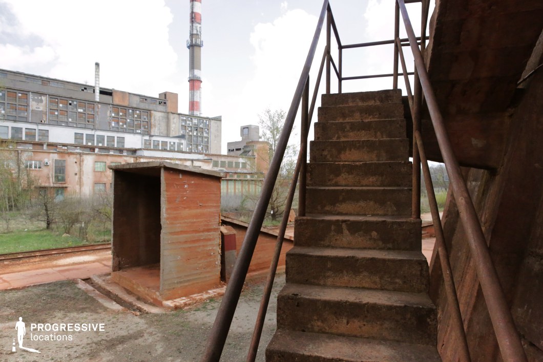 A rusted metal staircase with corroded railings leads up to an elevated industrial platform, set against a backdrop of a weathered factory complex with broken windows and a towering smokestack. The aged concrete steps and rust-stained surfaces emphasize the decay of the location, while the surrounding overgrown vegetation contrasts with the stark, utilitarian structures. The railway tracks and remnants of metal structures suggest a formerly active industrial site now left to deteriorate. This location is ideal for filming post-apocalyptic, dystopian, or crime thriller scenes, offering an atmosphere of desolation, urban decay, and industrial abandonment.