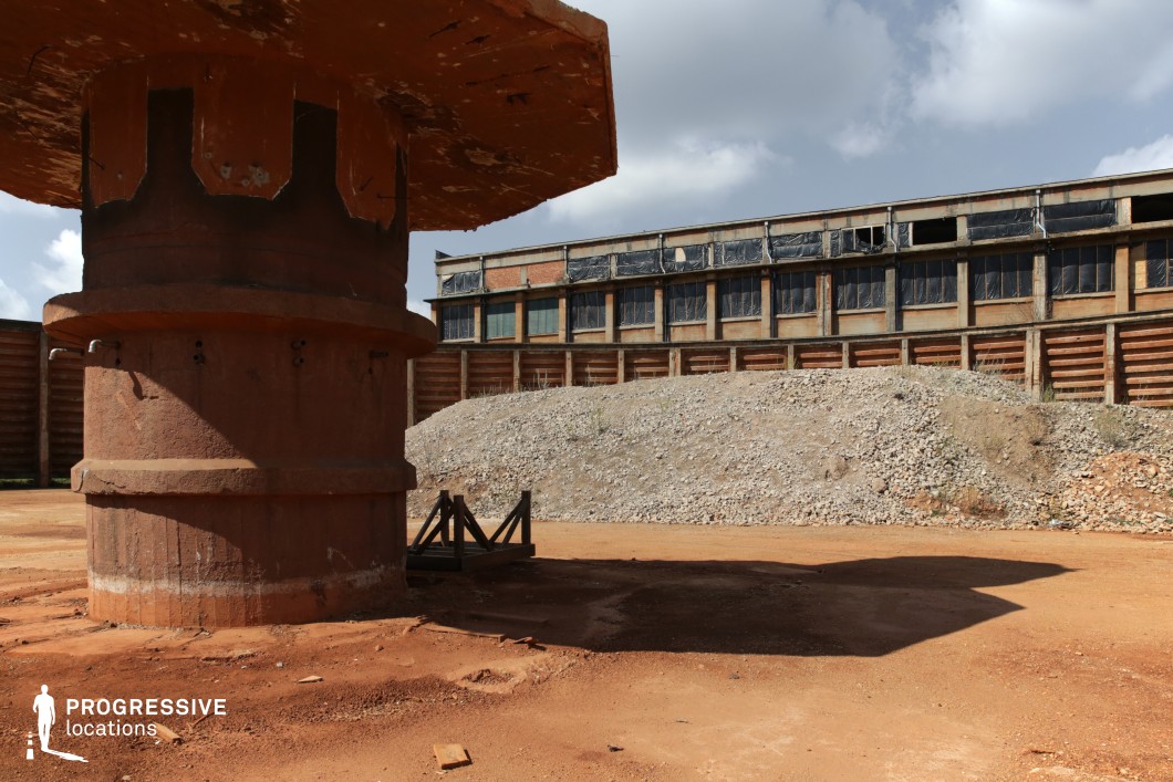This desolate industrial landscape features a large rusted cylindrical structure in the foreground, possibly a former machine base or storage component, with weathered bolts and corroded metal detailing. The ground is covered in reddish dirt and scattered debris, adding to the abandoned and rugged atmosphere. In the background, a long, deteriorating warehouse with broken windows and partially covered openings stretches along a raised gravel mound, bordered by a reinforced retaining wall. The combination of exposed concrete, rust, and decay creates a stark, post-apocalyptic setting. This location is ideal for dystopian, action, or thriller films, serving as a hideout, a battleground, or an abandoned research facility. The vast open space and industrial relics provide strong visual contrast for dramatic wide shots and intense chase sequences.