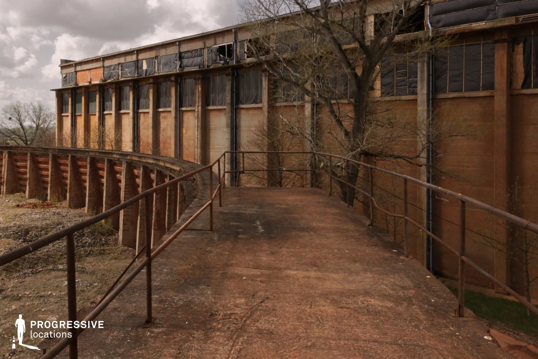 This desolate industrial location features a rusted elevated walkway with a corroded metal railing, leading towards a large, decaying factory building. The concrete structure is weathered and stained, with broken or missing windows, tattered coverings, and exposed framework adding to its abandoned aesthetic. The curved wall below follows the same industrial style, reinforcing the sense of scale and functional purpose of the site. Overgrown trees and encroaching vegetation further emphasize the site's long-term neglect. This setting is ideal for filming dystopian, post-apocalyptic, or crime thriller scenes, offering an atmospheric backdrop for narratives involving secrecy, danger, or forgotten histories. The combination of industrial decay and natural overgrowth creates a visually compelling contrast suited for eerie or suspenseful storytelling.