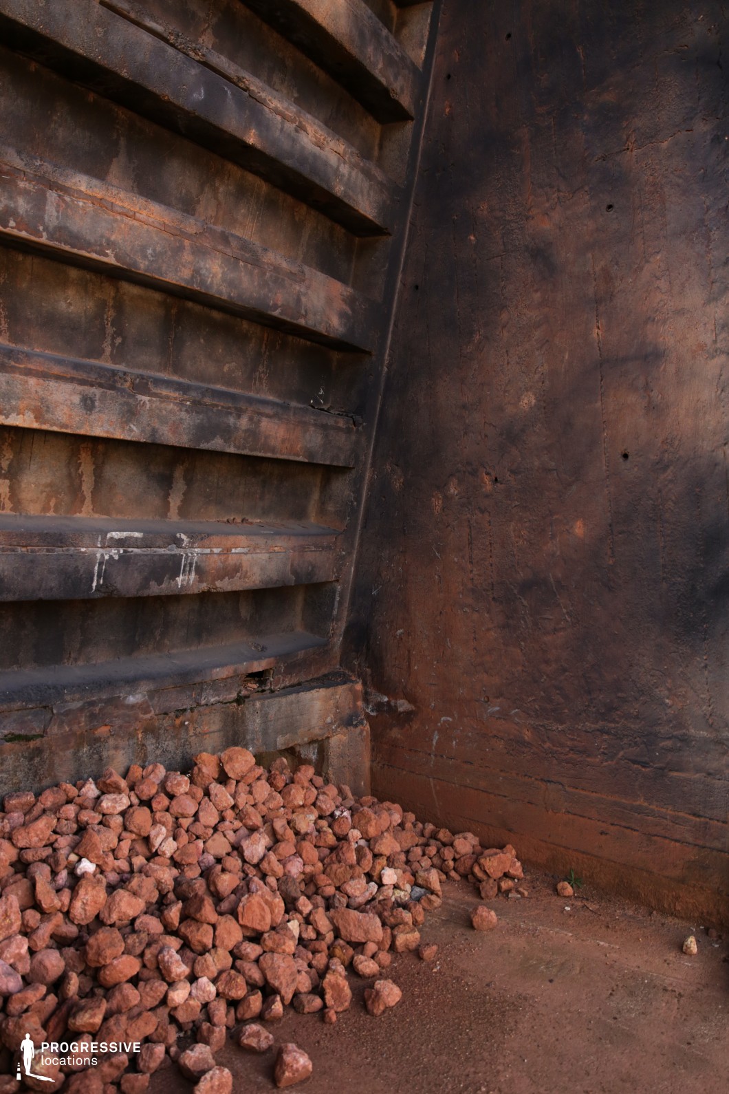 This gritty industrial setting features a heavily rusted metal structure with deep, weathered textures, conveying a sense of age and decay. The corroded steel panels, streaked with oxidation, contrast with the rough, reddish-brown rock fragments scattered at the base, suggesting a mining or construction environment. The muted, earthy color palette enhances the raw, industrial atmosphere, while the structural angles and riveted surfaces add depth and ruggedness to the composition. This location is ideal for filming dystopian, post-apocalyptic, or crime-related scenes, providing a visually striking backdrop for intense, high-stakes storytelling. The enclosed space and worn-down materials make it perfect for sequences involving confinement, tension, or clandestine industrial operations.