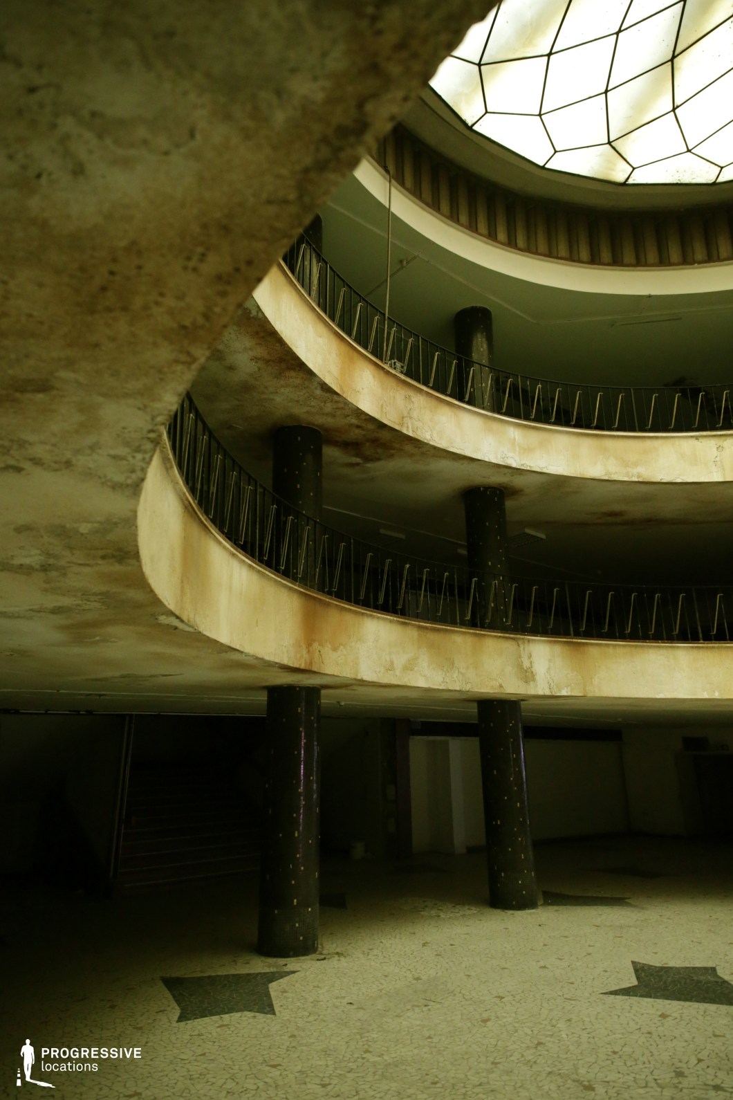 This decayed grand hall features a multi-level circular design with curved balconies supported by dark, mosaic-clad columns. The railings have a mid-century modern aesthetic, while the stained and peeling surfaces reveal years of neglect. A large geometric skylight filters soft, diffused light into the space, emphasizing its former grandeur. The terrazzo floor, decorated with star motifs, hints at a past function related to entertainment or public gatherings. This setting is ideal for filming haunting, atmospheric scenes in psychological thrillers, dystopian films, or noir-inspired narratives, providing a visually rich, mysterious backdrop for intrigue, secrecy, or lost grandeur.