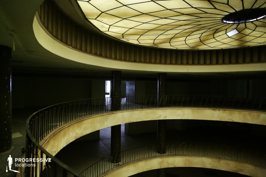 The image depicts a hauntingly atmospheric, multi-level atrium with curved walkways and a striking geometric skylight. The aged, yellow-tinted ceiling panels cast a dim, diffused light, enhancing the space’s eerie and forgotten ambiance. The peeling paint, rusted railings, and darkened corners emphasize its state of abandonment. Vertical columns add to the symmetry, while open doorways in the background suggest an expansive, empty interior. This location is ideal for filming psychological thrillers, dystopian narratives, or neo-noir films, offering a surreal, almost hypnotic visual setting for scenes involving intrigue, suspense, or exploration of the past.
