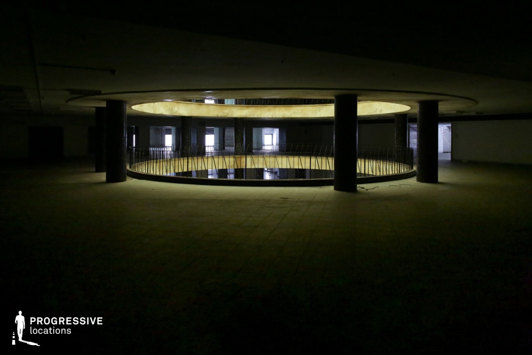 This dimly lit, cavernous interior features a striking circular atrium, supported by thick black columns and lined with a deteriorating metal railing. The layered architectural design, with concentric openings between floors, creates a dramatic and almost surreal visual depth. Sparse lighting casts eerie shadows, enhancing the sense of abandonment and decay. The open doorways in the background suggest a once-functional but now desolate space, ideal for filming psychological thrillers, dystopian sci-fi, or neo-noir narratives. This location provides an unsettling yet visually compelling backdrop for clandestine meetings, chase sequences, or moments of eerie solitude.