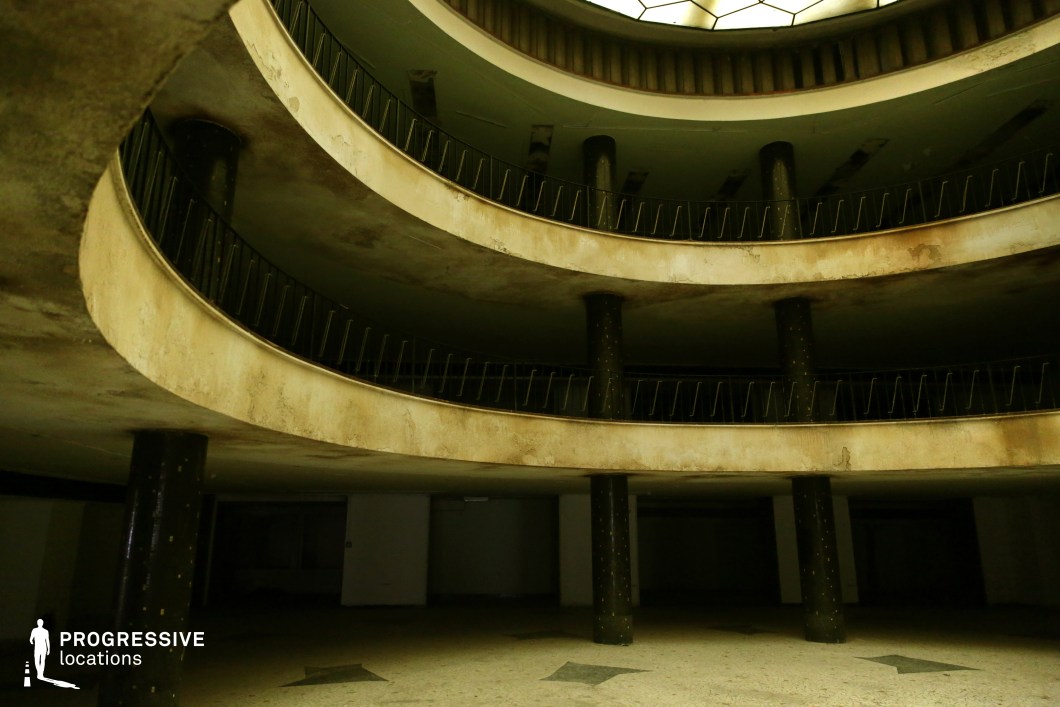 This hauntingly atmospheric space features a multi-level circular atrium with curved balconies that encircle the central area. The worn, stained concrete and dark, cylindrical columns give the location a sense of decay and abandonment, while the dim lighting from above creates a moody, almost surreal ambiance. The repetitive geometric patterns in the railings and the layered architecture add visual depth, making it a striking backdrop. The space is ideal for filming dystopian, sci-fi, or psychological thriller scenes, evoking a sense of mystery, secrecy, or past grandeur now lost. The eerie stillness and shadowy recesses make it especially suited for unsettling or suspense-driven sequences.