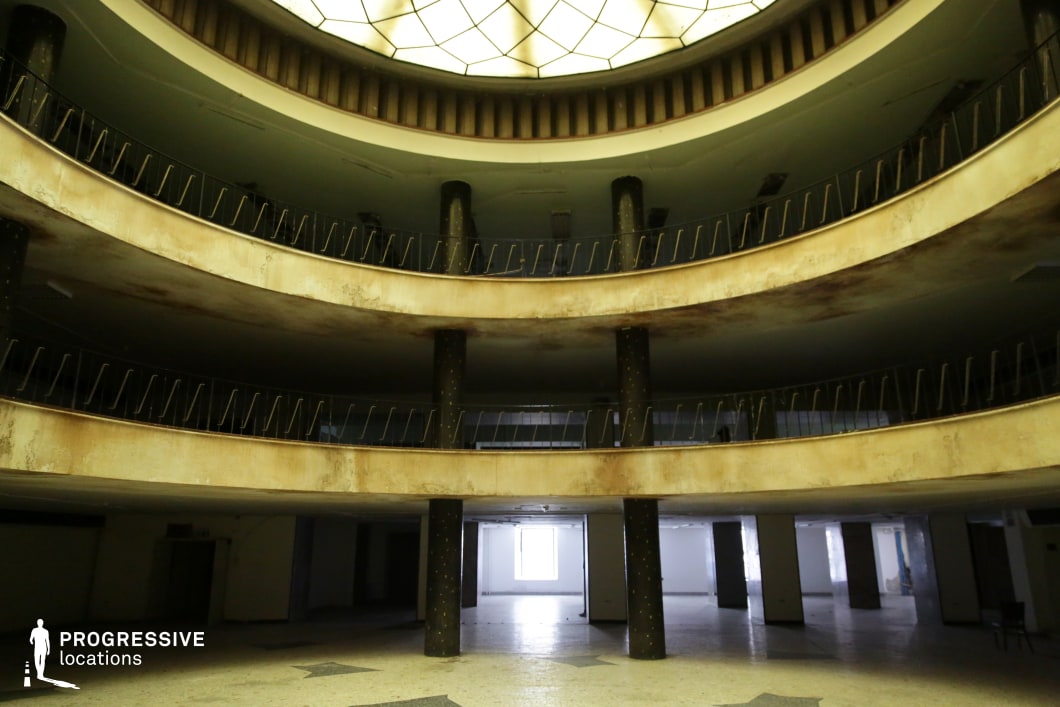 This eerie, multi-level atrium features a symmetrical, curved structure with rust-streaked balconies, black cylindrical support columns, and a striking stained-glass skylight that casts a dim, greenish glow over the decayed interior. The peeling surfaces, empty corridors, and broken railings add to the sense of abandonment, evoking a once-grand public space, possibly a former theater, hotel lobby, or government building. The geometric repetition and vast open floor create a haunting yet cinematic atmosphere. This location is ideal for filming psychological thrillers, dystopian sci-fi, or neo-noir films, serving as a backdrop for unsettling encounters, clandestine meetings, or explorations of a lost, forgotten world.
