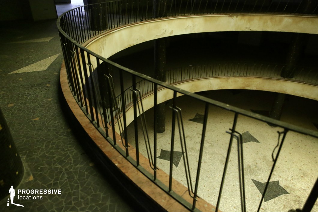 This abandoned circular balcony structure features an aged metal railing with a repeating linear pattern, enclosing a multi-level open space. The terrazzo flooring is adorned with faded black star motifs, adding a nostalgic and cinematic quality. The peeling paint on the curved walls and the worn mosaic tiles on the surrounding floor contribute to the location’s vintage, slightly eerie atmosphere. The curved architectural elements and soft, dim lighting create a sense of depth and movement. This space is ideal for noir, mystery, or psychological thriller productions, serving as a setting for clandestine meetings, hidden histories, or dramatic revelations in a forgotten theater, former hotel lobby, or underground venue.
