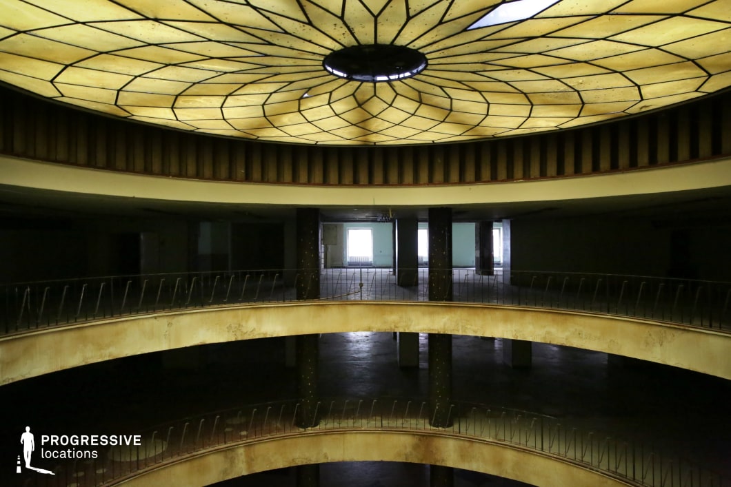 The image captures a grand, abandoned atrium featuring a multi-level circular balcony and a striking stained-glass ceiling. The intricate geometric dome structure diffuses a yellowish glow, creating a surreal and atmospheric effect. The balconies, lined with rusted railings, show signs of wear, with peeling paint and weathered surfaces adding to the aged, forgotten ambiance. The central open space below, along with the dimly lit, empty rooms in the background, reinforces the feeling of desolation. This location is ideal for filming eerie, suspenseful, or historical scenes, making it perfect for mystery, neo-noir, or psychological thriller genres. The dramatic lighting and decayed grandeur create a haunting yet visually compelling backdrop for narratives involving secrecy, abandonment, or rediscovery.