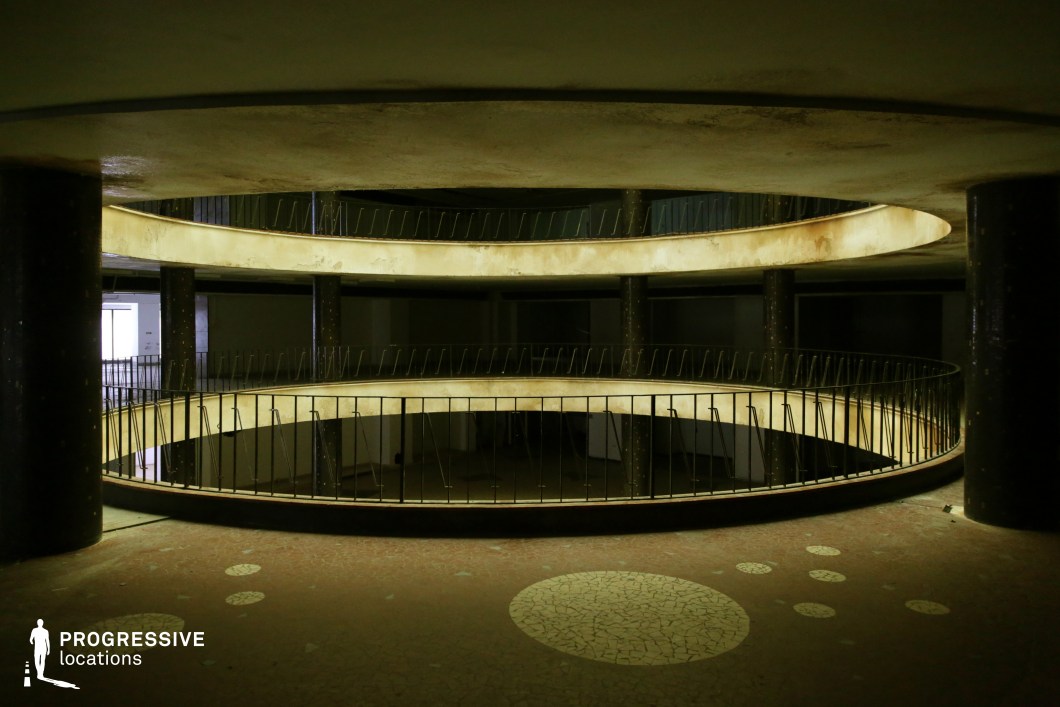 This image showcases a multi-level, circular atrium with decaying balconies and dark structural columns, evoking a sense of forgotten grandeur. The curved architecture, coupled with dim lighting and peeling surfaces, suggests a mid-century modern or socialist-era design now left in neglect. The railings feature thin, rusted metal bars, adding to the eerie and desolate aesthetic. The terrazzo flooring with circular patterns contrasts with the surrounding decay, hinting at a past vision of modernism. This location is ideal for filming dystopian thrillers, psychological dramas, or surreal, dreamlike sequences, offering a haunting, cinematic space with strong visual depth and symmetry.