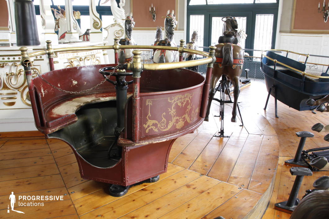 This beautifully preserved vintage carousel features intricately designed carriages and hand-painted wooden horses, evoking the charm of early amusement parks. The rich wooden flooring and decorative gold accents contrast with the aged patina on the ride’s mechanisms, enhancing its nostalgic appeal. The detailed craftsmanship, including elegant scrollwork and brass railings, suggests a historical fairground attraction or a turn-of-the-century entertainment piece. This location is ideal for filming whimsical, nostalgic, or eerie sequences in period dramas, fantasy films, or psychological thrillers, where the setting adds depth to themes of innocence, memory, or mystery.