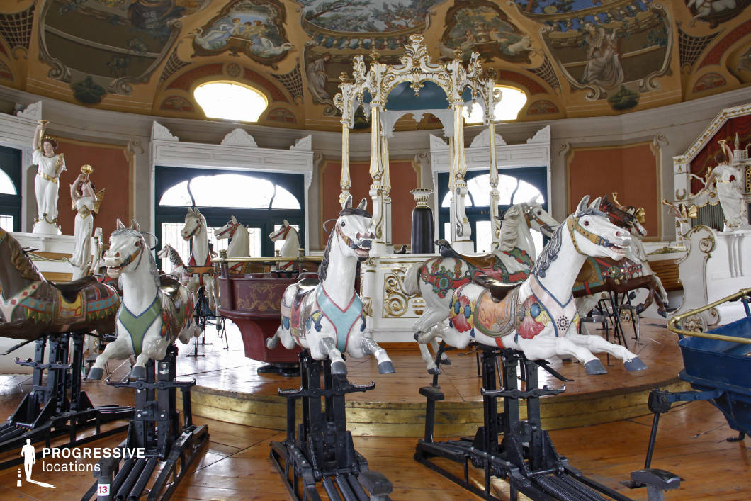 This opulent vintage carousel, set within an elaborately decorated indoor space, features meticulously crafted and colorfully painted horses mounted on mechanical bases. The richly detailed ceiling, adorned with classical frescoes, gold embellishments, and sculpted figures, enhances the whimsical and nostalgic atmosphere. The warm wooden flooring and soft natural light filtering through arched windows contribute to the timeless elegance of the scene. This location is ideal for filming fantasy, period dramas, or psychological thrillers, offering a surreal yet nostalgic backdrop for dreamlike sequences, romantic encounters, or eerie moments of suspense in a forgotten fairground.