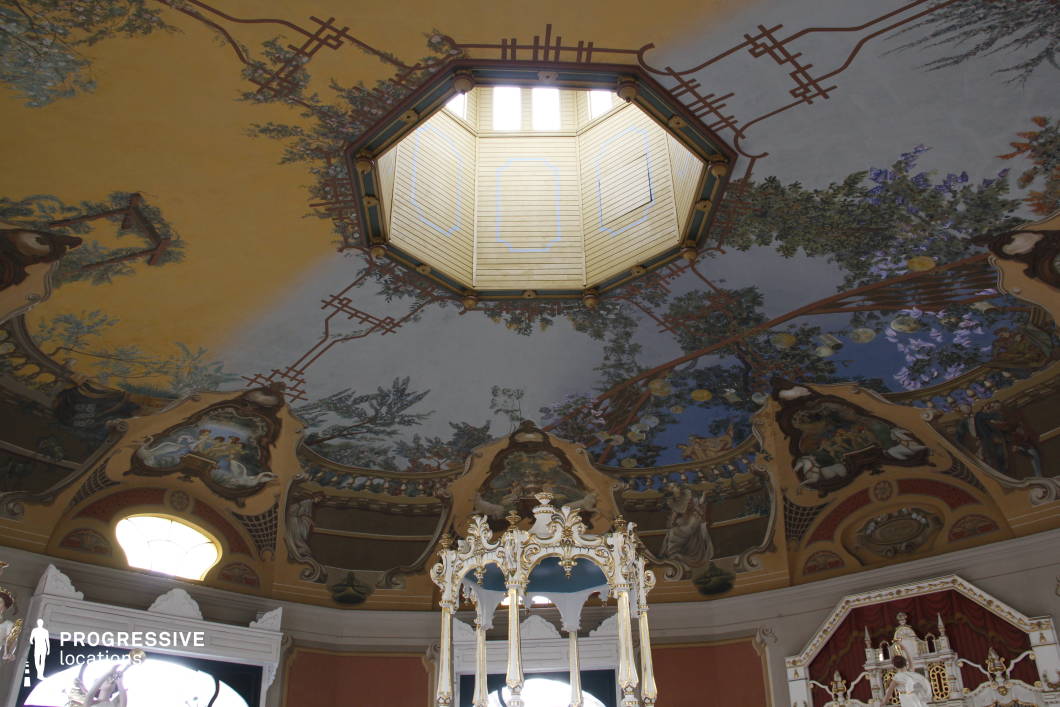 This opulent ceiling showcases an elaborate fresco adorned with nature-inspired motifs, delicate floral patterns, and classical figures, all radiating from an octagonal skylight. The intricate artwork, rich in pastel hues and gold detailing, evokes a grand, historical ambiance reminiscent of Baroque or Rococo architectural styles. The interplay of light filtering through the skylight enhances the depth and vibrancy of the frescoes, while the surrounding sculptural moldings and decorative elements contribute to the luxurious atmosphere.